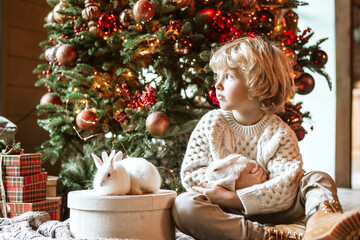 little kid boy with blond hair and blue eyes in beautiful knitted clothes sits in living room near Christmas tree in chalet with white small rabbits for family celebration of Christmas and New Year