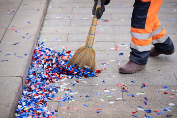 A janitor cleans up colorful confetti with a broom on the side of a city street after a celebration event - obrazy, fototapety, plakaty
