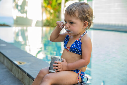 little girl eats yogurt by pool in swimsuit