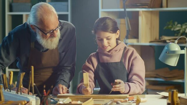 Grandfather Carpenter with his Teenage Granddaughter Works in the Workshop, Making Artistic Woodcarving. Communication of Generations. Handicraft, Skills Training. Profession, Art and Hobby Concept.