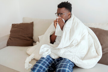 Sick man with cold sitting on bed and blow nose. Man feeling cold, sitting on bed and blowing his nose. Cropped shot of young man suffering with flu while sitting wrapped in duvet on the sofa at home.