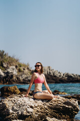 Happy young woman enjoying freedom on vacation while sitting on the coast rocks enjoying sea and blue sky view