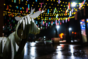 Footage of evening public park decorated for the celebrating winter holidays with metal deer in the foreground. Garlands and ribbons are hanging on an alley and light of lamps is reflecting on asphalt
