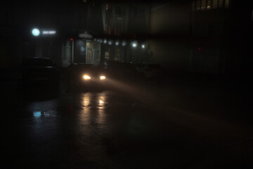 Car with shining headlights is standing on the parking next to shop in the residential district at foggy night. Automobile is on the dark smog street. Empty bedroom community at evening time in autumn