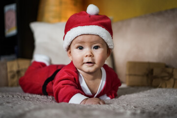 cute little newborn baby in christmas outfit with santa claus hat crawling on a couch in a cozy...
