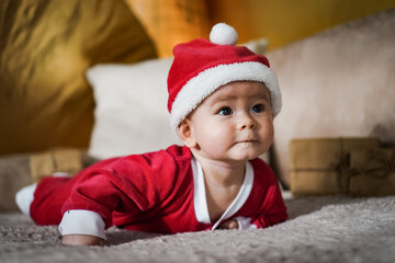 cute little newborn baby in christmas outfit with santa claus hat crawling on a couch in a cozy...