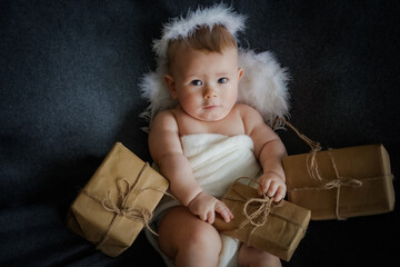 happy innocent newborn baby in white angel outfit with feather wings lying on a grey background with eco friendly natural paper gift boxes