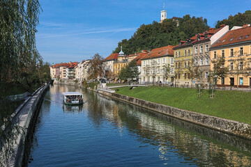Fototapeta na wymiar Ljubljana, Slovenia. Historical buildings along the Ljubljanica River and Castle Hill with lookout tower of Ljubljana Castle in sunny autumn day. View from the St. James's Bridge across the river.