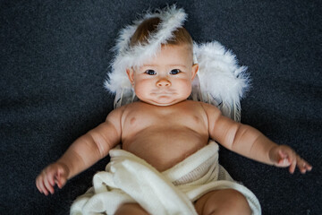 cute happy innocent newborn baby in white angel outfit with feather wings lying on a grey...