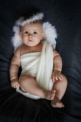 cute happy innocent newborn baby in white angel outfit with feather wings lying on a grey...