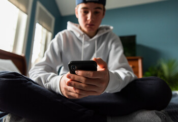 Low angle of teen boy in white sweater on smartphone in bedroom.