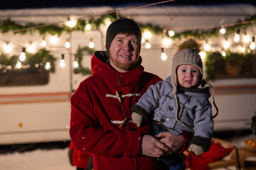 Caucasian red-haired man with a boy in his arms at the mobile home. Father and son celebrate Christmas on a trip.