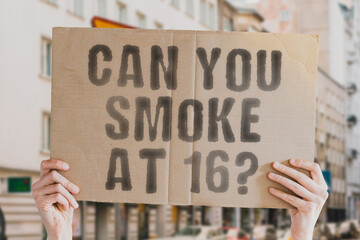 The question " Can you smoke at 16? " on a banner in men's hand with blurred background. Relationship. Control. Parents. Law. Illegal. Unlawful. Rule. Policy
