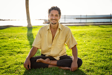 Smiling hippie italian man sitting in lotus, practicing yoga outdoor on sunny day. Men looking at the camera with cute smile