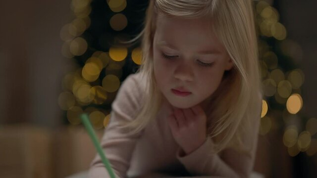Little Girl Of Four Years With Blond Long Hair. Behind It Christmas Tree Sparkles With Garlands. Girl Draws. She Is On Floor Near Christmas Tree.