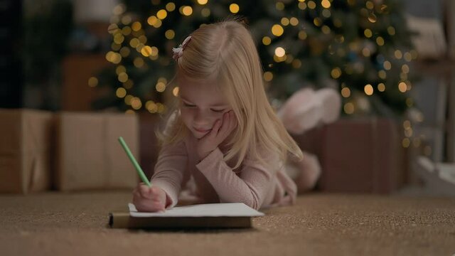 Little Girl Of Four Years With Blond Long Hair. Behind It Christmas Tree Sparkles With Garlands. Girl Draws. She Is On Floor Near Christmas Tree. Near Christmas Tree Gifts. She Smiles.