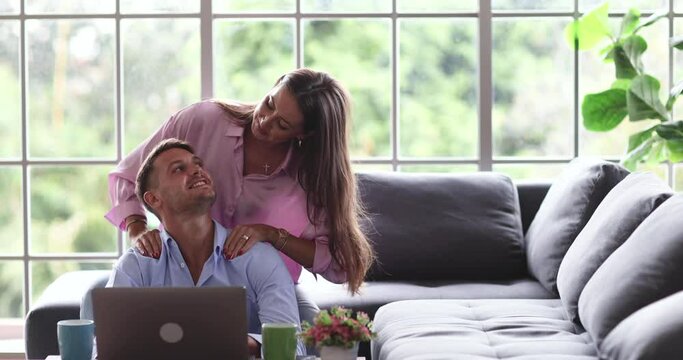 Millennial Caucasian beautiful lovely sweet housewife sitting on cozy sofa massaging her happy husband shoulder and arms while complimenting typing working with laptop computer on table in apartment.