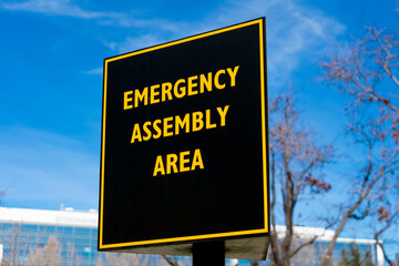 Emergency Assembly Area outdoor sign. Background commercial office building under blue sky.