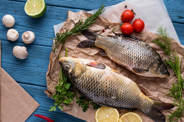 Top view two carp fish are lying on paper on a wooden blue background, next to ingredients, tomatoes, lemons and spices