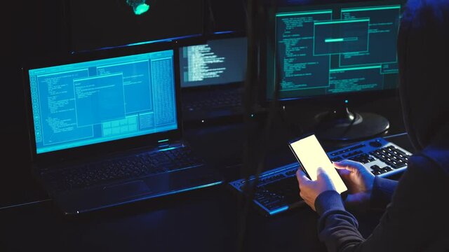 Overhead hacker in hood working at laptop and mobile phone typing text in dark room, An anonymous hacker uses malware with mobile phone to hack password, personal data steals money from bank. cyber