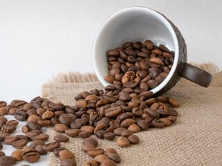 The coffee cup lies sideways on the table; grains of roasted aromatic coffee pour out of it onto a canvas napkin. Breakfast. Lunch break. Coffee business concept