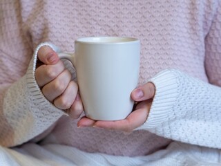 Female hands hold a white mug with cappuccino coffee. He clasps a mug with his palms, warms his hands in cold weather. A girl in a knitted light sweater, her legs are covered with a knitted blanket