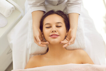 Happy young woman relaxing on massage bed or table at spa center, getting face massage, and enjoying rejuvenating facial treatment for bright complexion, even tone and clear fresh perfect glowing skin