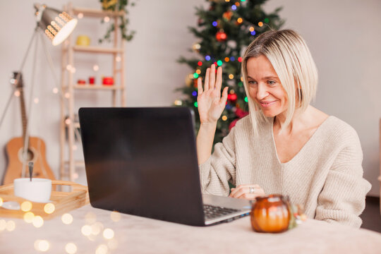 Excited adult 40s woman waving hello with hand, using laptop at home