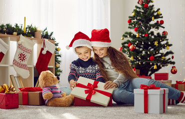 Obraz na płótnie Canvas Magic time. Cute happy kids boy and girl in Santa hats open box with Christmas presents on festive morning. Sister and brother open gift box sitting on warm floor in room with Christmas decorations.