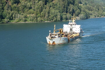 View on dredger ship from a merchant container vessel sailing through Columbia river, Oregon to direction Pacific Ocean. In background is coniferous forest with trees.