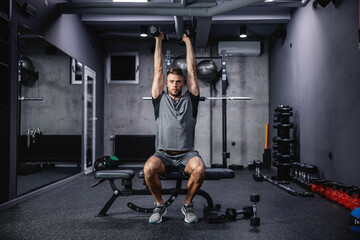 Fototapeta na wymiar Slim and fit. Static weight loss training for a slim and fitness body. A sportsman focused on training sits on a bench in the gym and raises his dumbbells above his head. Fitness lifestyle, sport