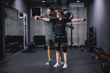 Fototapeta na wymiar Young woman workout in electric muscular suit, stimulate exercise at training center. A male trainer helps her improve her body position. A young woman in an EMS suit holds dumbbells in her hands