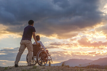 Woman Sitting in wheelchair and her care helper walking on mountain at sunset background.