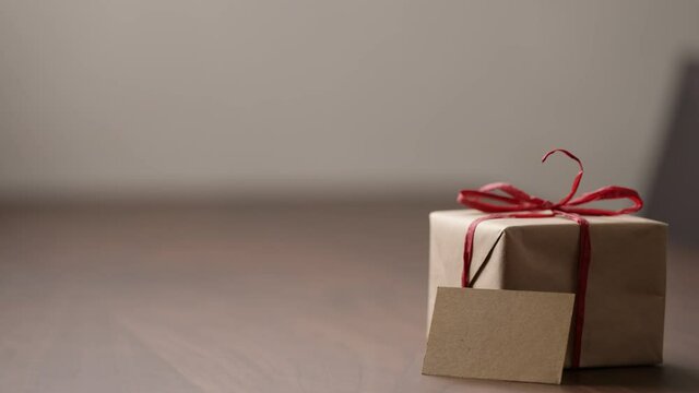 Slide tracking shot of brown paper gift box with red bow on wood table with blank paper card