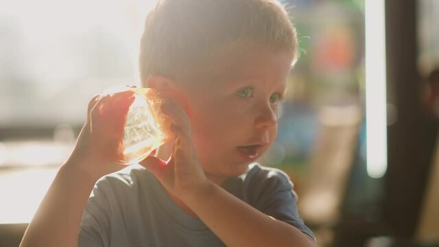 Curious little boy puts empty glass to ear and listens sea sounds having fun in room lit by sunlight at home extreme closeup slow motion