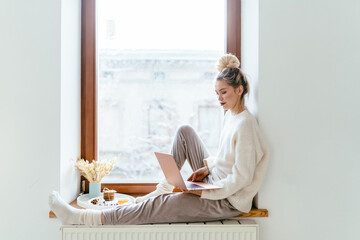 Young blond woman freelancer in cozy knitted white sweater sitting on windowsill with laptop computer working from home. Winter holiday mood. Enjoy sun in cold season.