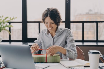 Young happy european woman female employee sitting at workplace in office and opening gift box