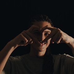 An adult woman with braided hairs,  piercing, the woman makes a fingers heart.