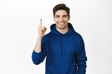 Fototapeta premium Smiling young man in hoodie, pointing finger up, showing advertisement, sale on top, standing over white background