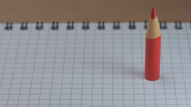 A small sharpened red wooden pencil appears and stands on an open checkered notebook, video clip, close-up