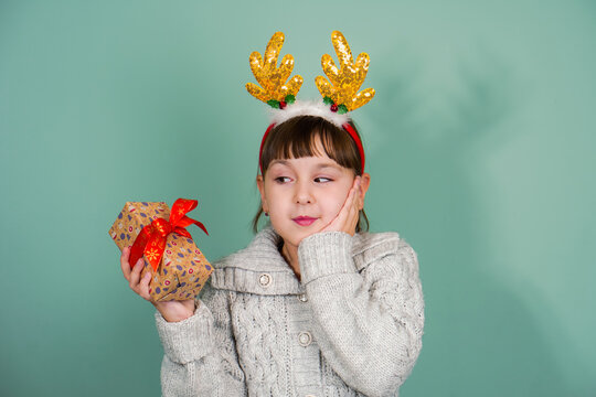 Studio Portrait Of Suprised Child Girl Wearing Yellow Deer Horns With Gift Box In Hand On Mint Background