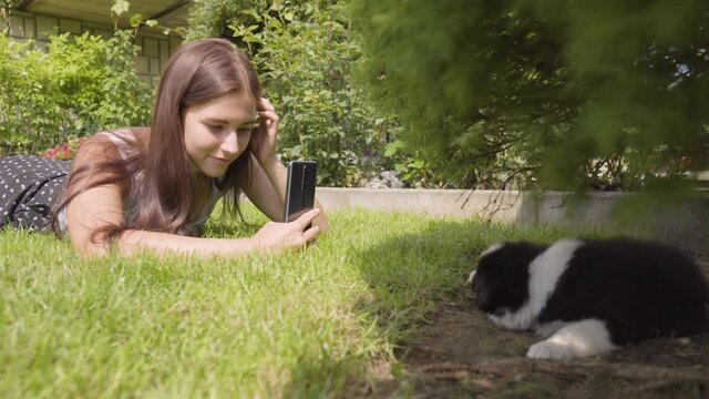 A young beautiful woman lies on grass and takes photos of a cute little puppy under a tree