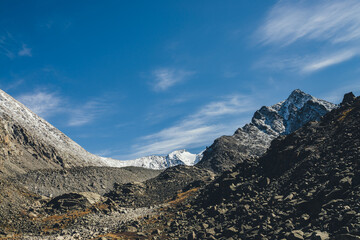 Awesome alpine landscape with dark rockies and high snow-covered mountains in sunshine under blue sky with cirrus clouds. Beautiful highland scenery with sharp black rocks and mountain top with snow.