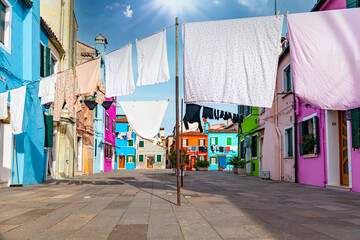 Many clothes hanging out to dry in a village of very colorful and cheerful houses on a sunny day. Clothes hanging outdoors in a small square on the island of Burano, Venice, Italy. - obrazy, fototapety, plakaty
