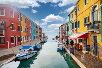 Fototapeta na wymiar Burano Island, Venice, Italy. View of a canal in the town of Burano with the brightly colored houses famous all over the world. There are colored boats in the canal. Sunny blue sky with clouds.