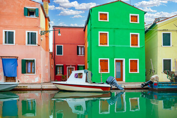 Fototapeta na wymiar Burano Island, Venice, Italy. View of a canal in the town of Burano with the brightly colored houses famous all over the world. There are colored boats in the canal. Sunny blue sky with clouds.