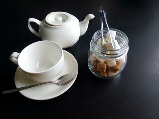 A white cup and saucer, a jar of sugar and a teapot, isolated on a black background