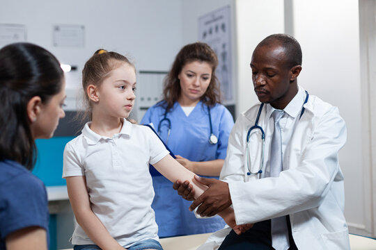 African american pediatrician doctor bandage fractured bone of girl patient during clinical physiotherapy in hospital office. Young child having broken arm after accident. Health care service