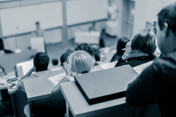 Speaker giving presentation in lecture hall at university. Participants listening to lecture and making notes.