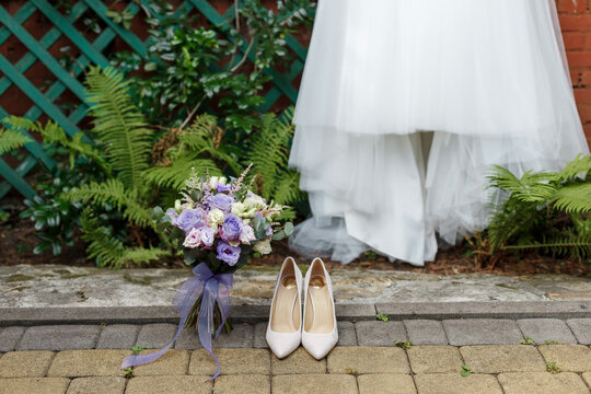 Wedding details: classic bride shoes and bouquet of roses standing on the background pf dress and greenary. Very Peri - trendy color of the year 2022 in weddings flowers. 
